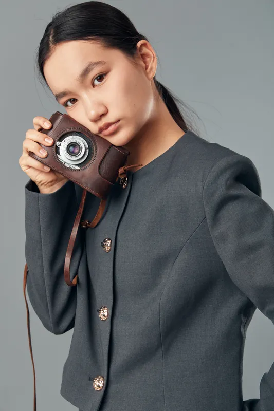 Young woman holding vintage camera near face wearing grey buttoned jacket with serious expression on neutral background studio portrait fashion photography model.