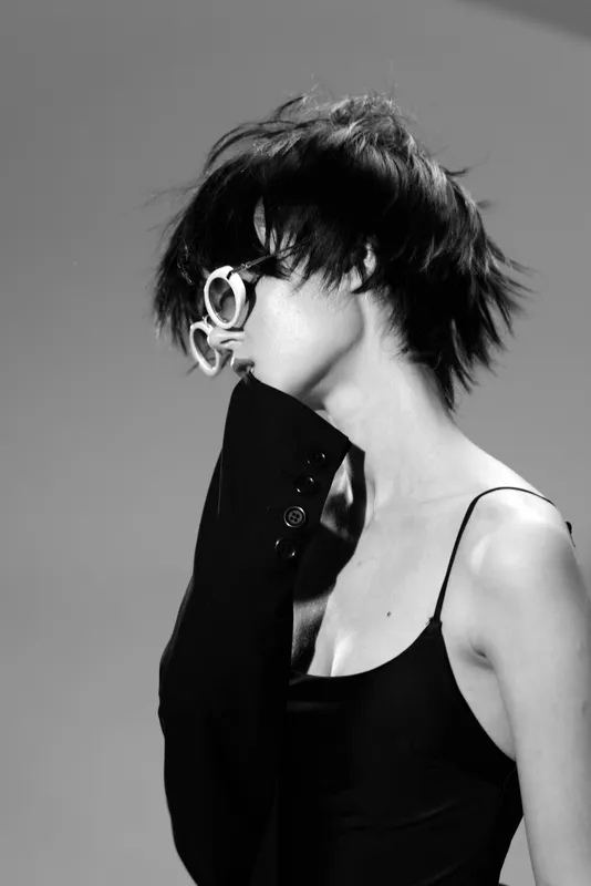 Black and white portrait of a stylish woman with short hair wearing sunglasses and a black outfit in a minimalistic setting