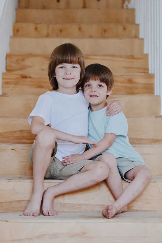 Two cute boys sitting on wooden stairs, sharing a moment of warmth and affection. The natural light enhances the cozy atmosphere, perfect for family themes.