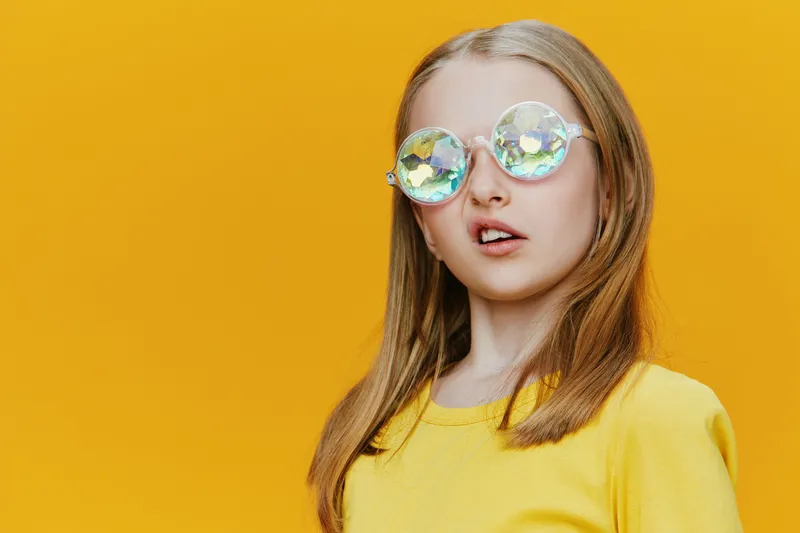 A joyful young girl wearing oversized holographic sunglasses and a bright yellow shirt, surrounded by a vibrant yellow background, exuding happiness and playfulness