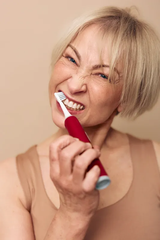 Mature woman with short blonde hair brushes her teeth aggressively with an electric toothbrush. Close-up on neutral background. Concept of dental care and oral hygiene routine.