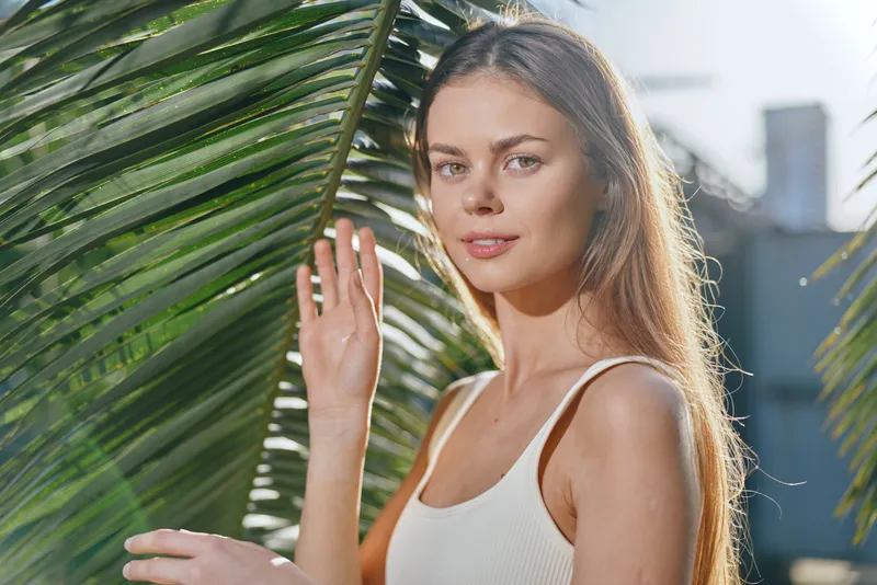 Young woman smiling outdoors by green palm leaves wearing a white tank top in natural sunlight with a clear sky background.