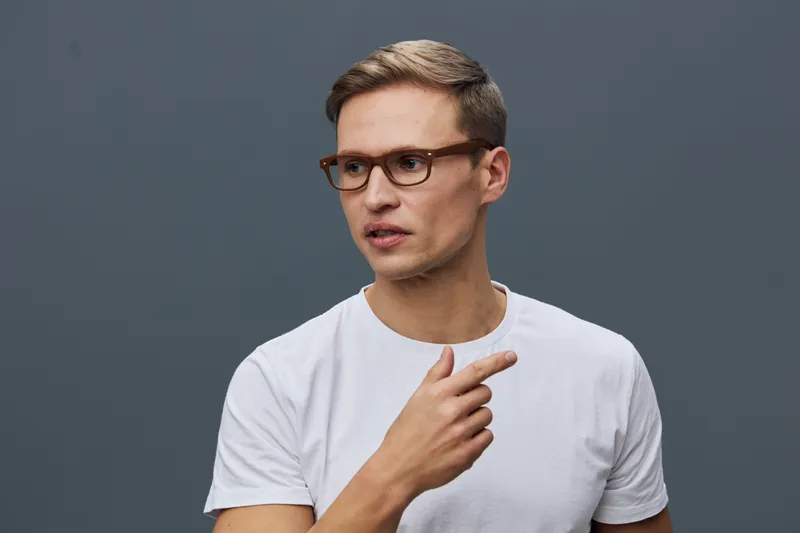 Thoughtful young man with glasses wearing casual white t-shirt on gray background, expressing curiosity or pondering topic, indoor studio portrait, people concept, modern style, lifestyle shot.