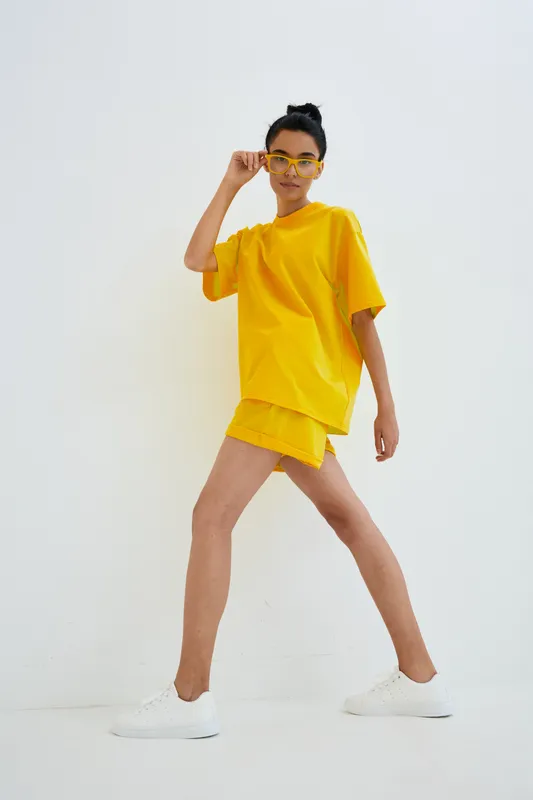 young woman in bright yellow oversized T-shirt and shorts posing confidently with sunglasses and casual white sneakers on white minimalistic background