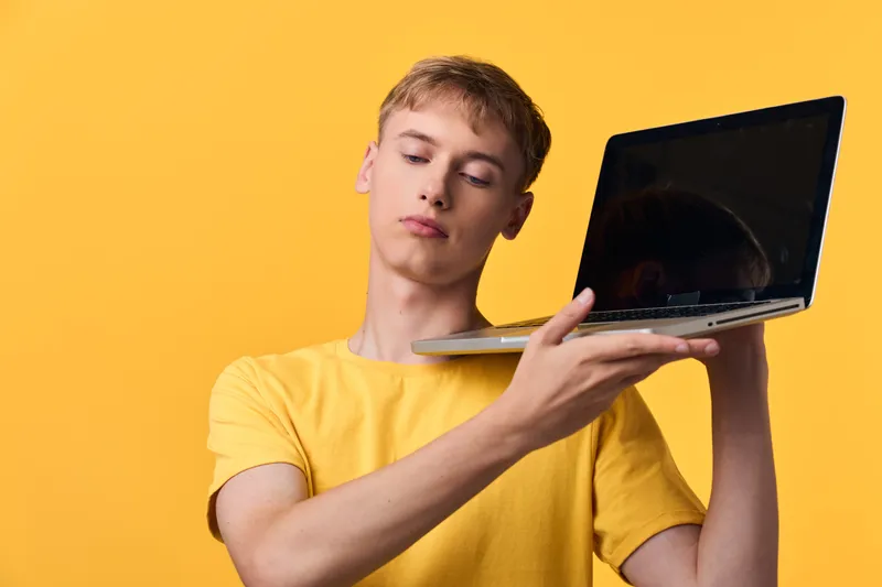 A yellow shirted man lifts a laptop against a vivid yellow backdrop, highlighting technology use, portability, and a casual workspace vibe with a modern device and bright color.