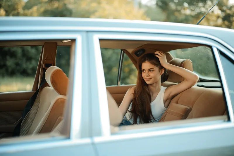 Young woman enjoying a sunny day inside a vintage car, wearing a white tank top, surrounded by lush greenery, reflecting a carefree summer vibe.