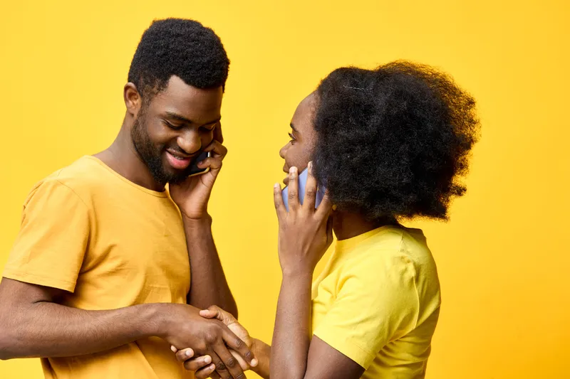 Cheerful African couple communicating on mobile phones, wearing casual yellow outfits against a vibrant yellow background, conveying joy and connection