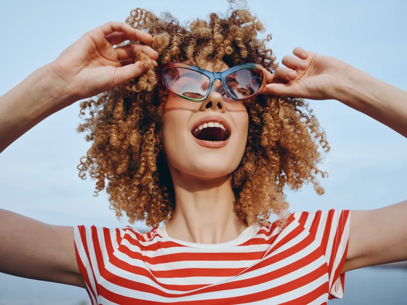 Joyful person with curly hair, oversized sunglasses, and a red striped shirt, expressing carefree summer energy under a bright blue sky and sunny breeze.