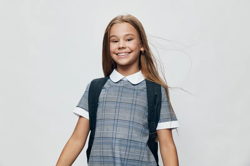 Girl with backpack smiling and standing isolated on white background, wearing checked dress and white shirt, happy child ready for school or education in studio.