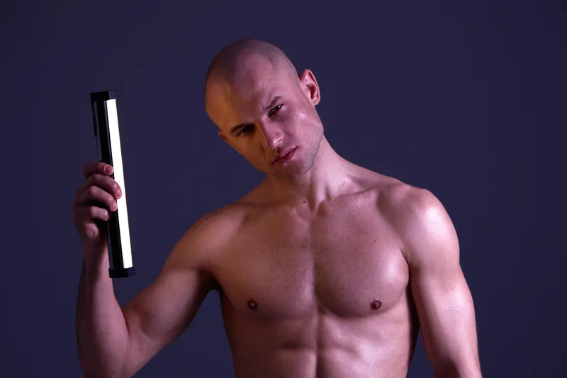 A confident man in a studio setting, holding a light bar, showcasing strength and style. The image has vibrant colors, capturing a modern aesthetic.