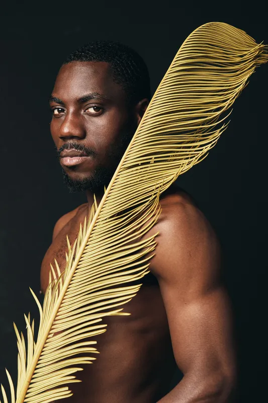 African man with dark skin and bare torso holding large yellow palm leaf in creative conceptual visual style on black background. Artistic portrait with natural texture and expression.