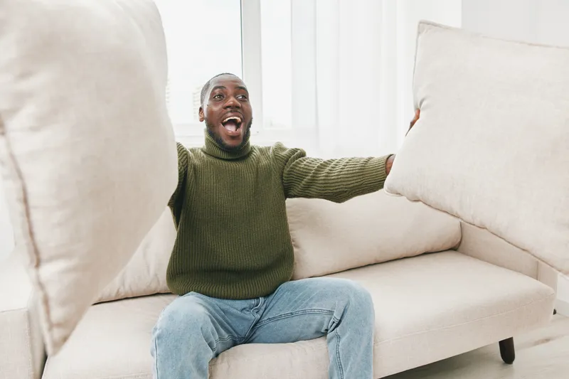 Happy young Black man celebrating at home with cushions, wearing a green sweater and jeans, joyful expression, bright modern interior design