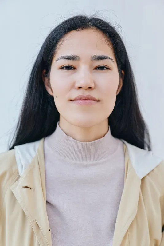 Young woman with a calm expression wearing a beige jacket over a pastel sweater in a light outdoor setting, simple casual styling, clean background, natural daylight, everyday fashion, relaxed