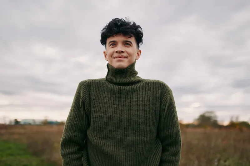 A young man in a green sweater stands outdoors with a calm smile, a natural portrait emphasizing authenticity and credibility through candid expression, unretouched details, and simple everyday
