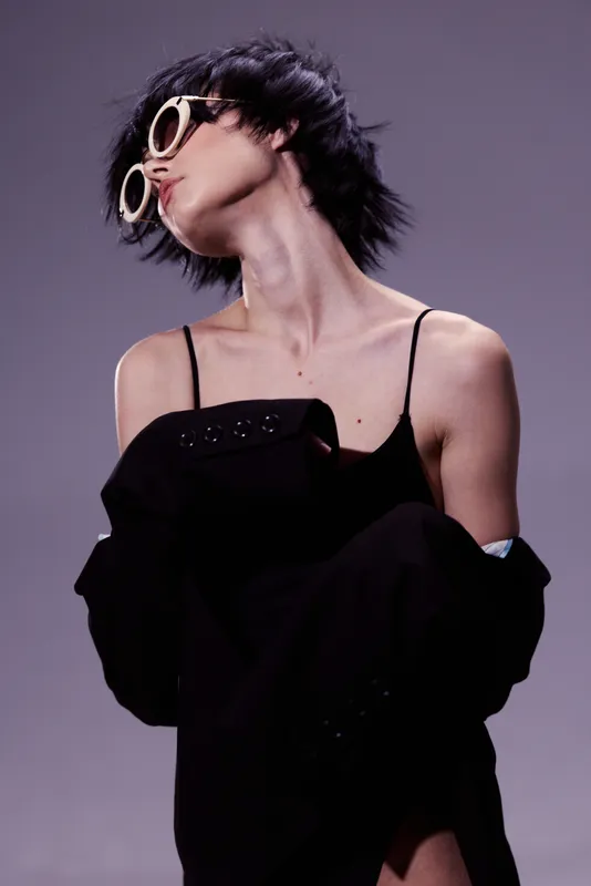 Stylish woman in sunglasses wearing black clothing with a modern short haircut posing confidently on a gray background.