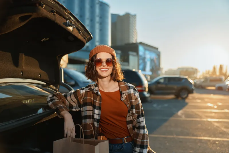 woman car shopping parking trunk smile sunglasses lifestyle: Young woman leans on car trunk with shopping bags, smiling in urban sunset. Authenticity, candid lifestyle, golden hour glow, mindful