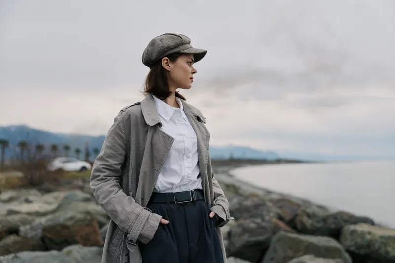 Young woman in stylish autumn outfit stands on rocky shore looking out at cloudy sky and distant mountains. Fashion concept with natural landscape and moody atmosphere outdoors.