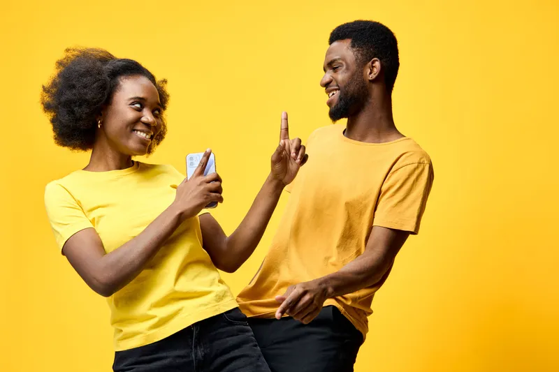 Joyful young African couple enjoying time together in bright yellow tops, using smartphone on vibrant yellow background, conveying happiness and togetherness