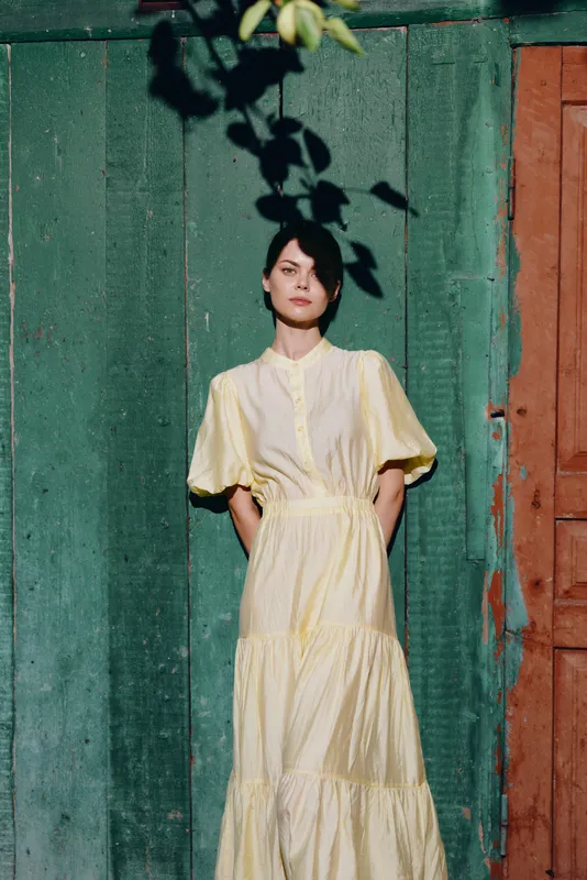Woman portrait in yellow dress standing by wooden wall in sunlight shadow