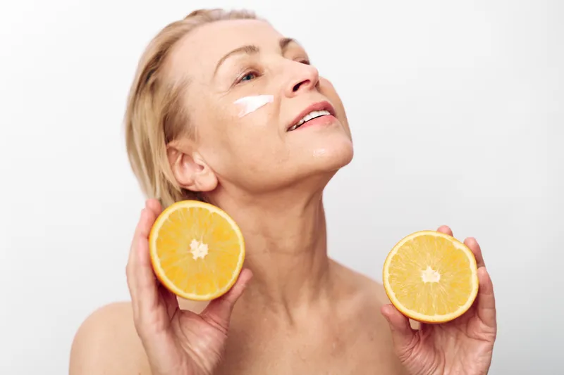 Mature woman with face cream on cheek holds two orange halves near face, smiling with closed eyes. Concept of natural skincare, healthy lifestyle, and citrus beauty treatment.