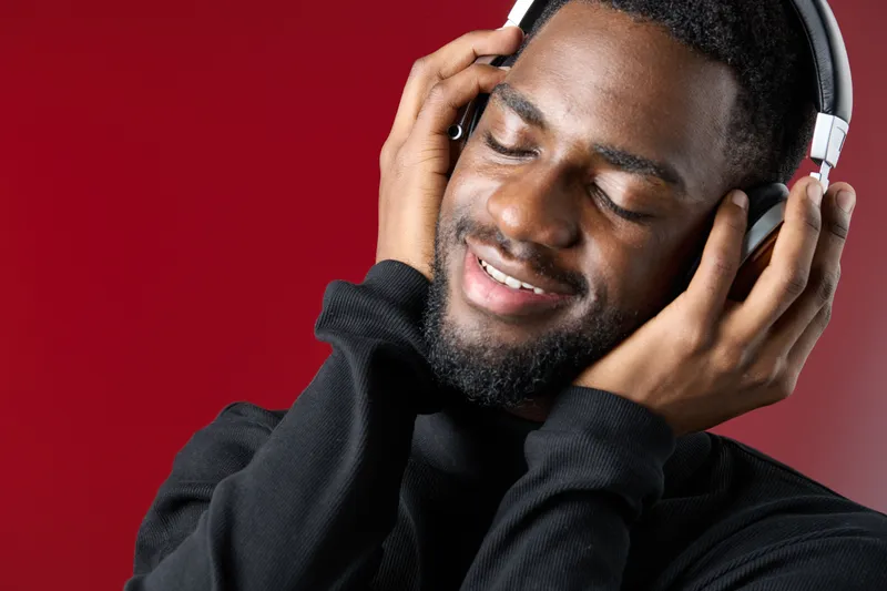 Young Black man enjoying music with headphones in a vibrant red background, expressing happiness and relaxation through his emotions