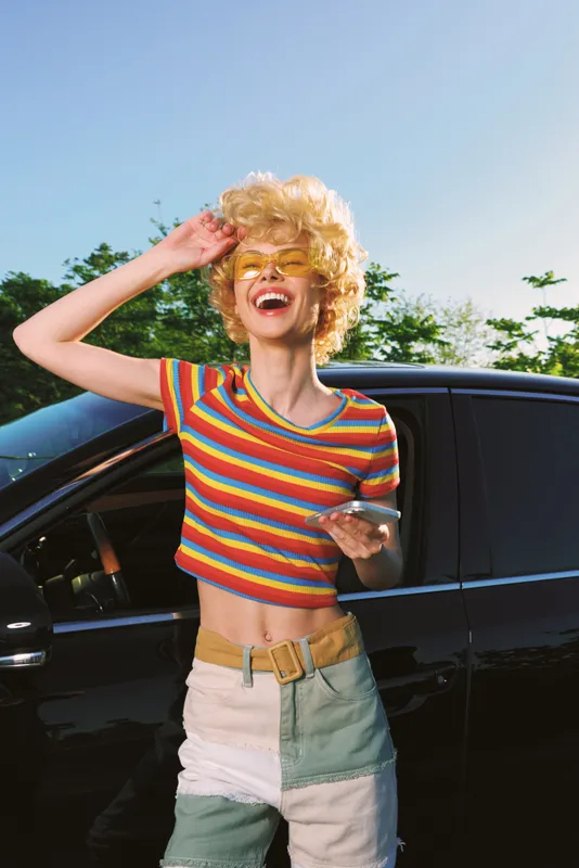 Joyful woman by a car, wearing a colorful striped shirt, embodies lifestyle and retro charm, outdoors under sunny sky, capturing candid, vibrant moment.