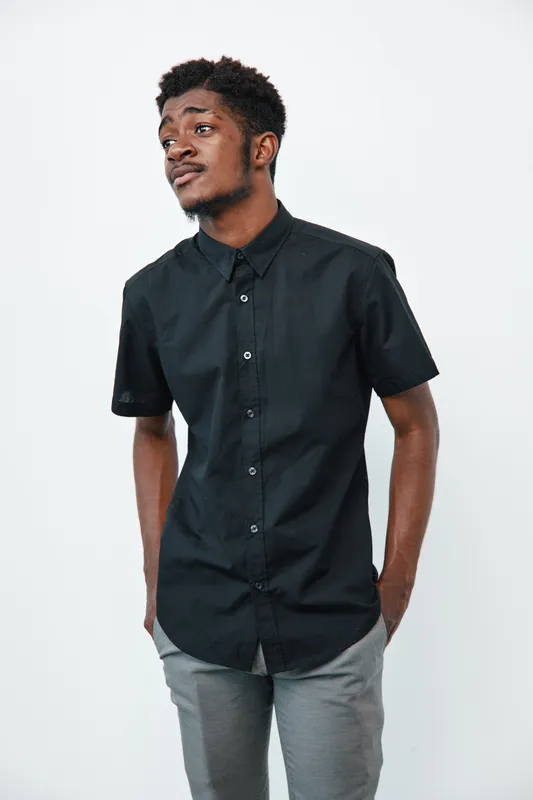 Young black man with dark skin wearing black short sleeve shirt and gray pants standing with hands in pockets on white background. Casual style and thoughtful expression.