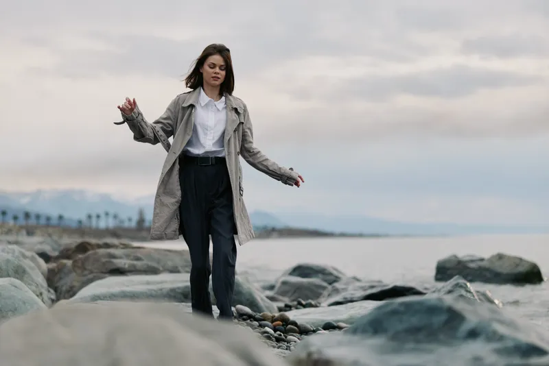 Young woman in a coat walks carefully on large rocks by the sea under cloudy sky. Concept of balance, nature, and outdoor calmness near water in cool weather conditions.