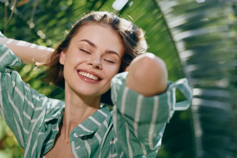 Joyful portrait of a smiling woman under bright sun, eyes closed, relaxed outdoors, surrounded by lush greenery and tropical light.