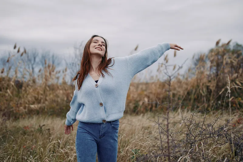An authentic candid portrait of a joyful woman in a natural field, arms outstretched, soft sunlight, casual sweater, conveying genuine warmth and everyday moments.