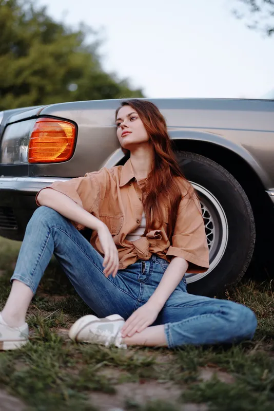 Young woman with long hair wearing a casual beige shirt and blue jeans, sitting beside a vintage car in a natural setting, showcasing a relaxed and stylish vibe.
