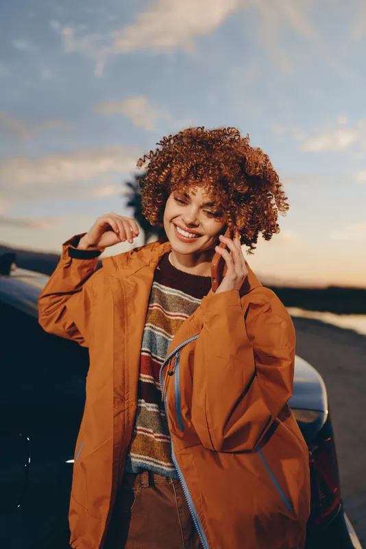 Woman lifestyle outdoors, smiling in warm sunset light, curly hair stirred by wind, adjusting her orange jacket, casual travel vibe, near sea horizon, golden evening sky