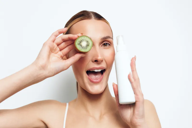 Young woman holding kiwi slice near eye and white cosmetic bottle, smiling with open mouth, clean skin, natural makeup, minimalistic background, beauty skincare concept.