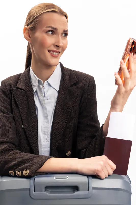Smiling woman in brown blazer holds passport and smartphone while sitting on suitcase. Concept of travel, vacation planning, and modern lifestyle with positive expression and casual outfit.