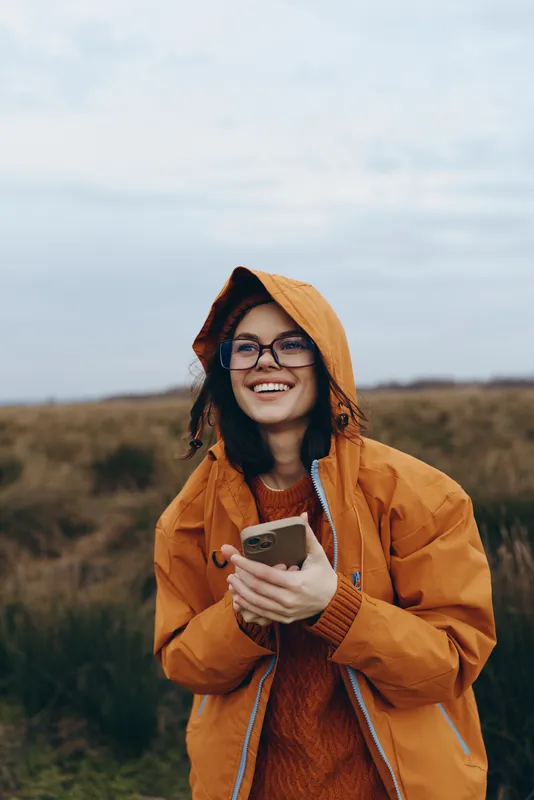 Woman lifestyle outdoors portrait, hooded raincoat orange, holding phone smiling, glasses visible, autumn countryside field background, casual travel mood, bright daylight, light wind
