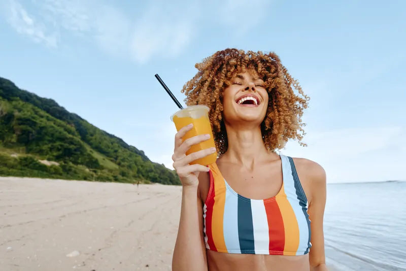 Woman on beach smiling and laughing, holding a takeaway drink cup with straw, sunny seaside background, curly hair and striped swimsuit, summer vacation mood, relaxed travel day
