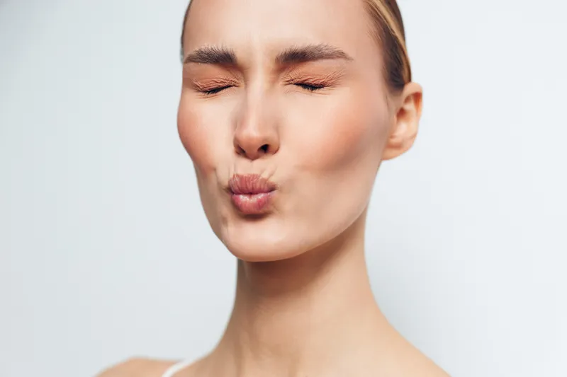 A young woman with closed eyes puckers her lips as if blowing a kiss against a plain light background. The image conveys playful, affectionate, and carefree emotions.