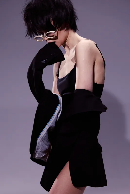 Stylish woman in black dress and jacket posing gracefully with glasses against a muted purple background