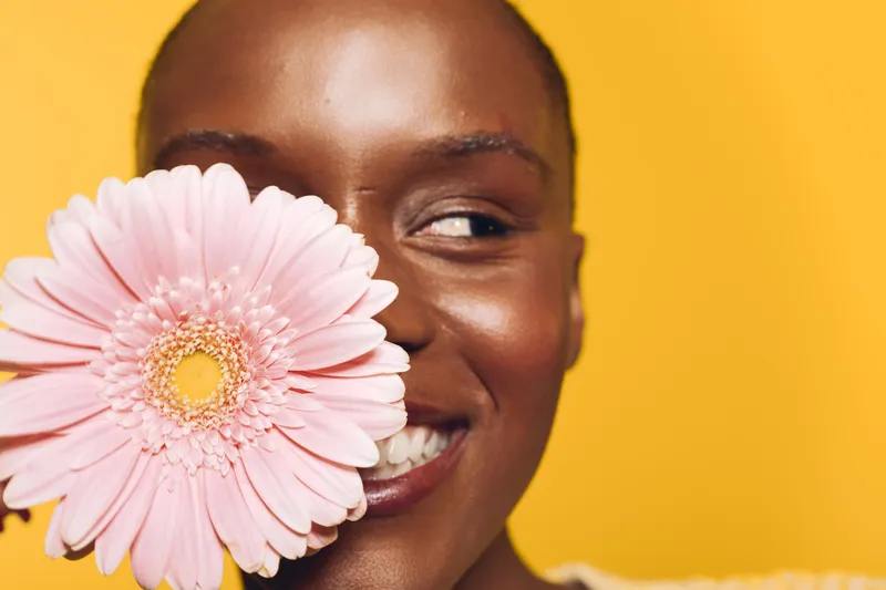 Close-up of smiling dark skinned African woman holding pink flower near face, looking sideways. Bright yellow background. Concept of beauty, happiness, natural skin and floral wellness.