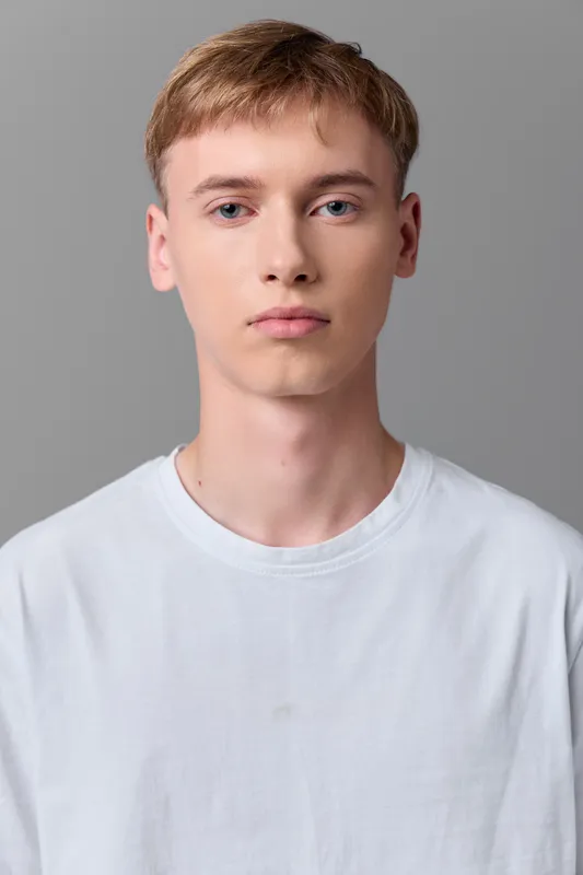 Young man with light skin and short hair in a white shirt stands against a gray backdrop, offering a calm, direct gaze ideal for stock imagery, profile headers, or fashion basics
