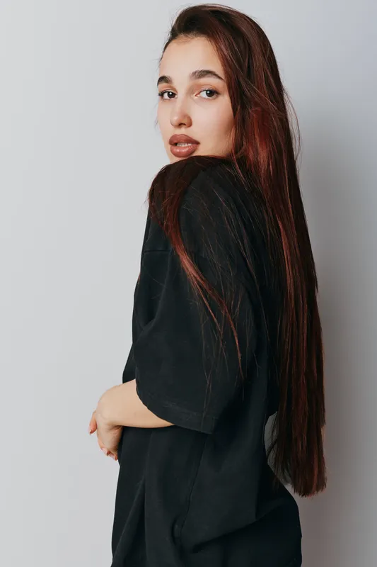 Young woman with long brown hair wearing black shirt posing sideways on gray background. Female model looking over shoulder with natural makeup and casual style in studio portrait.
