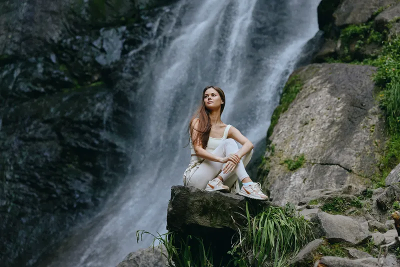 young woman sitting on rock near waterfall in forest, wearing casual white outfit and sneakers, enjoying nature and peaceful moment outdoors