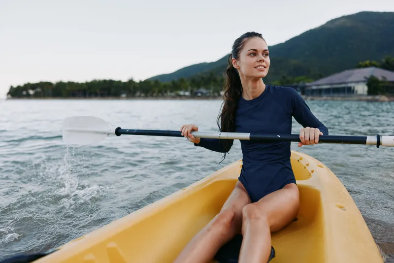A focused female paddler sits in a bright yellow kayak, gliding across calm water with a steady paddle stroke, mountains and distant shoreline framing the scene.