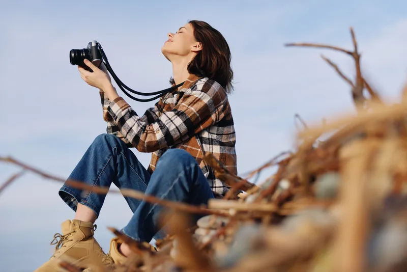 A photography of a woman in a plaid shirt and jeans sits on driftwood, camera raised toward the sky, capturing outdoor nature and a calm landscape moment with quiet focus