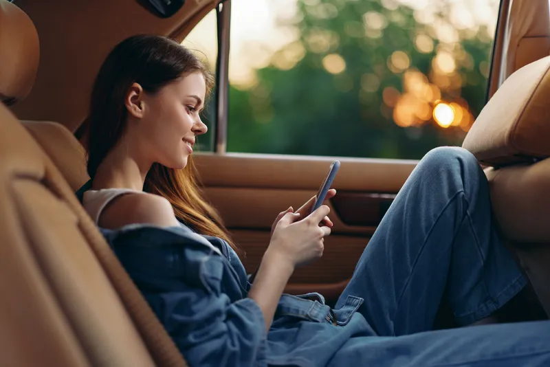 Young woman in casual denim attire relaxing in a car, using a smartphone against a warm sunset backdrop, embodying tranquility and modern lifestyle.