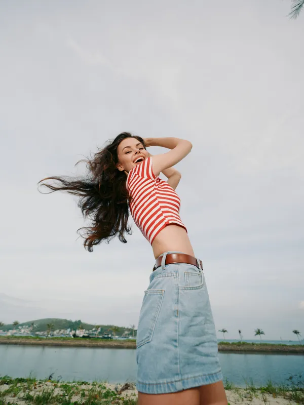 Young woman in a red and white striped top and denim skirt dances with wind in her hair beside a calm lake under a wide blue sky.