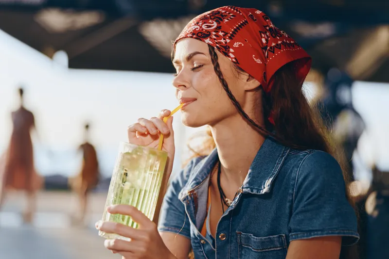 Lifestyle woman in modern boho-western denim street style sipping a refreshing drink outdoors, captured with warm film color tones for a relaxed summer vibe.