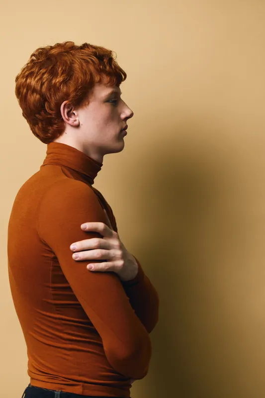 Young man with red hair wearing brown turtleneck stands in profile with arms crossed on beige background indoor studio portrait of confident thoughtful person.