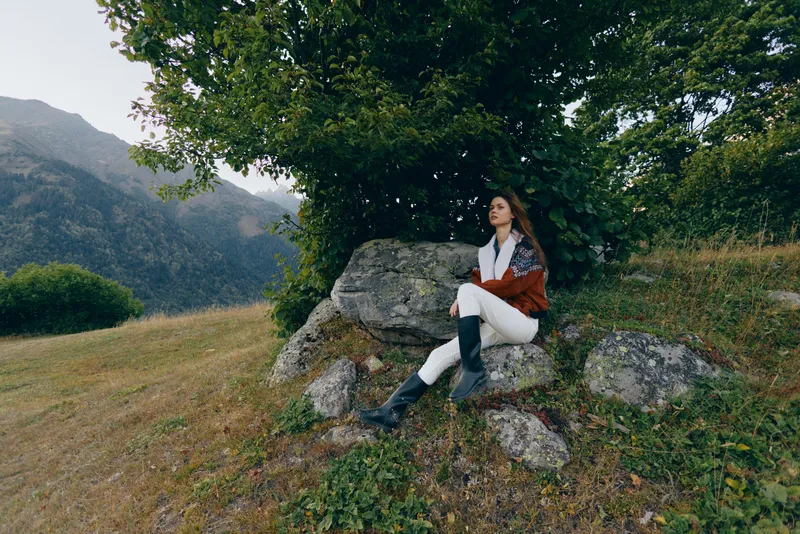 woman nature mountains tree rock meadow outdoors sitting on stone under leafy canopy, relaxed portrait in countryside with distant hills, scenic landscape and soft overcast sky