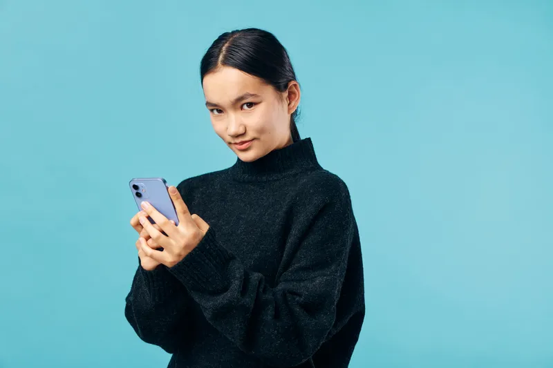 Woman standing against blue background holding smartphone texting with focus and slight smile wearing black sweater casual modern technology and communication concept